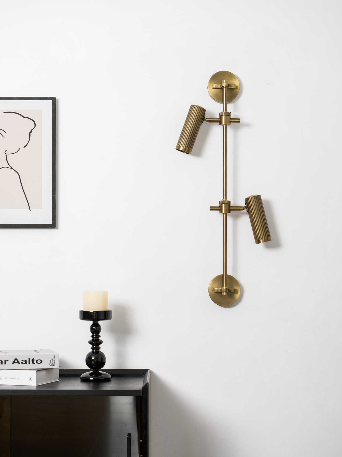 Bronze wall light with two heads from Clérachic, unlit, on a white wall above a dark sideboard with a black candlestick.