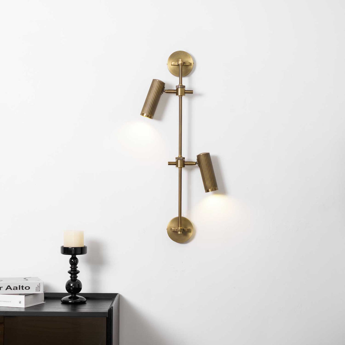 Two-head Clérachic wall light on a white wall, lit with a soft pool of light under each head, above a black candlestick.