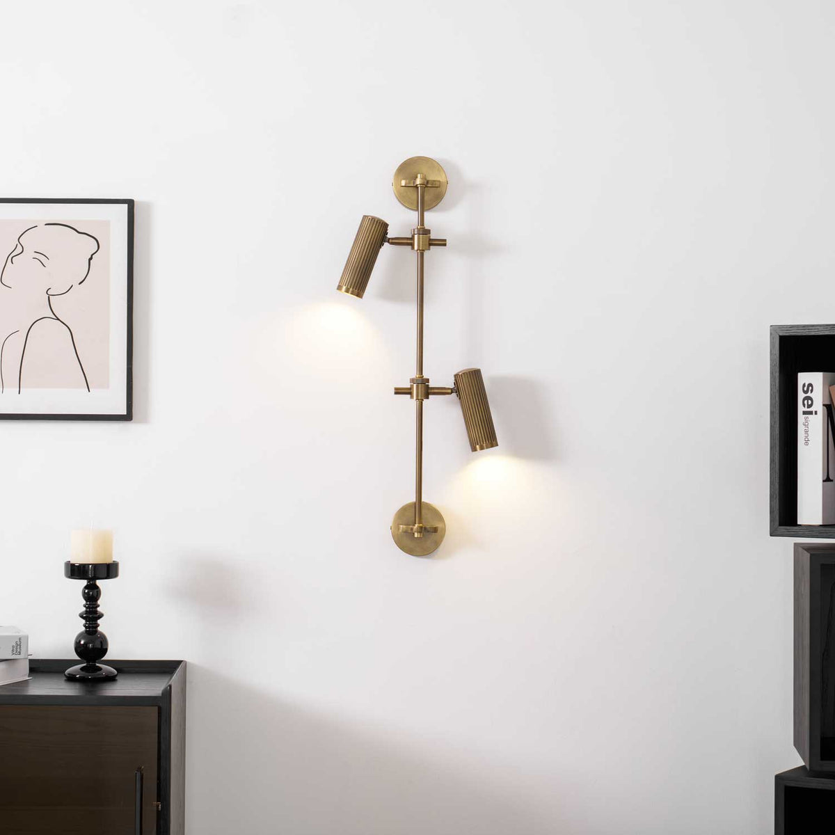 A lit two-head Clérachic wall light on a white wall, with a framed line drawing and a dark cabinet in the corner.