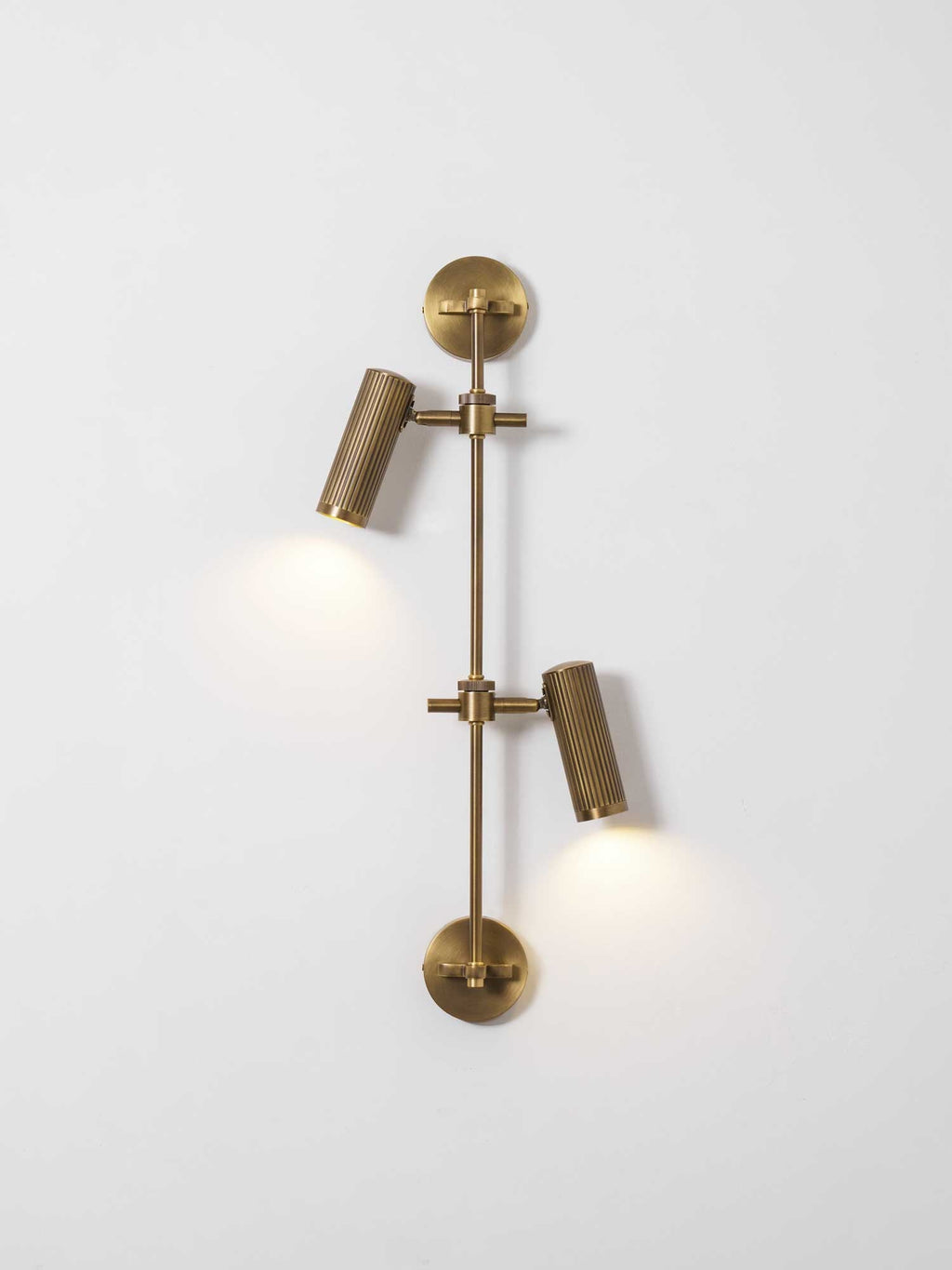 Lit two-head Clérachic wall light on a white wall, with a soft glow below each fluted bronze head.