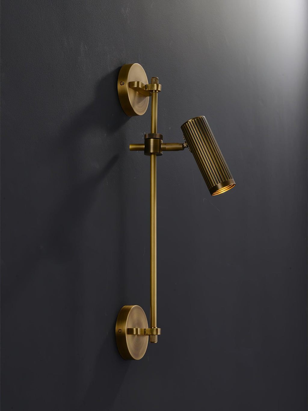 Single-head bronze wall light from Clérachic, unlit, on a dark charcoal wall with its round backplate at both ends of the rail.