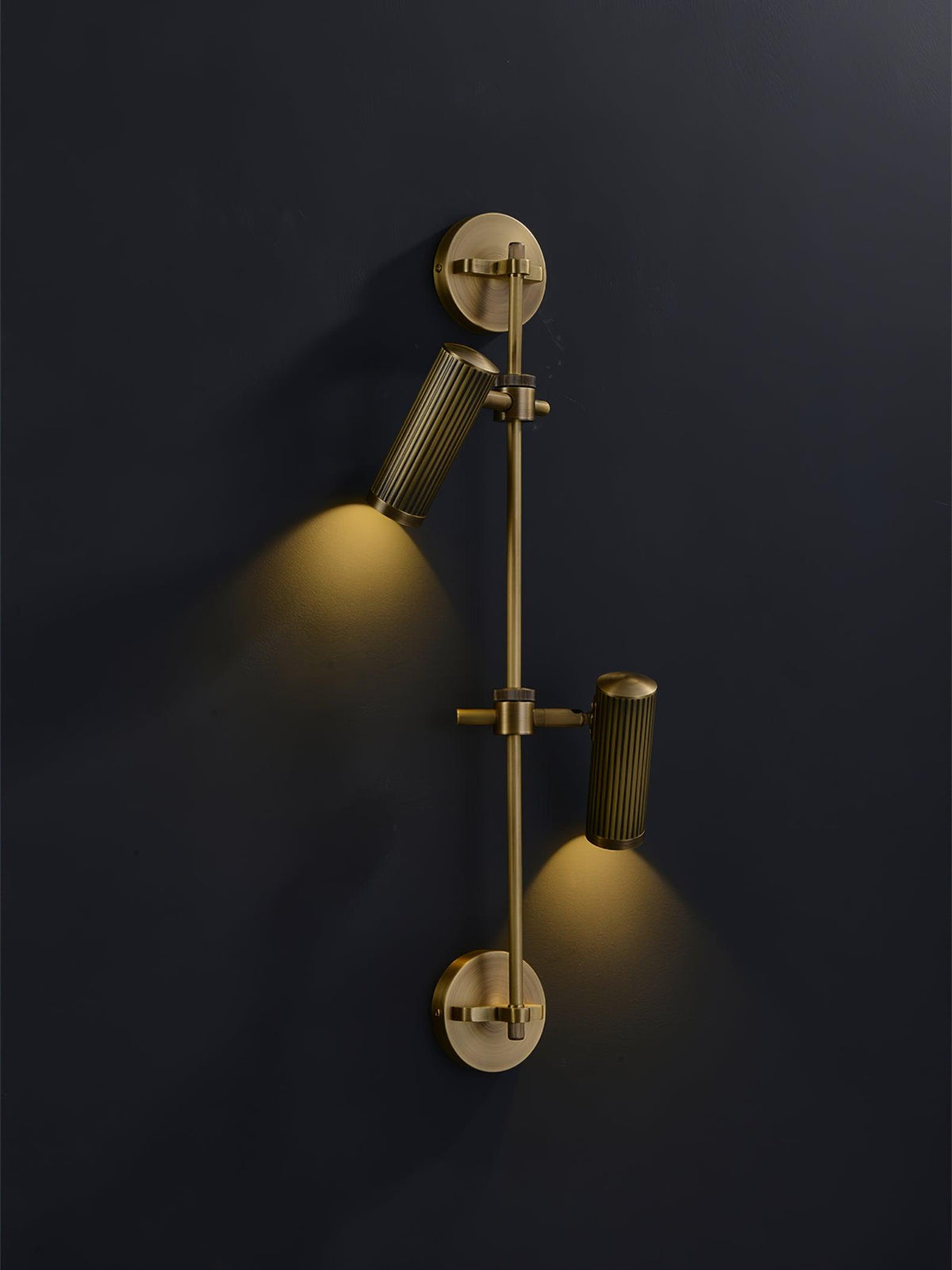 A lit two-head Clérachic wall light on a dark wall, with the upper head aimed down and a pool of warm light under each head.
