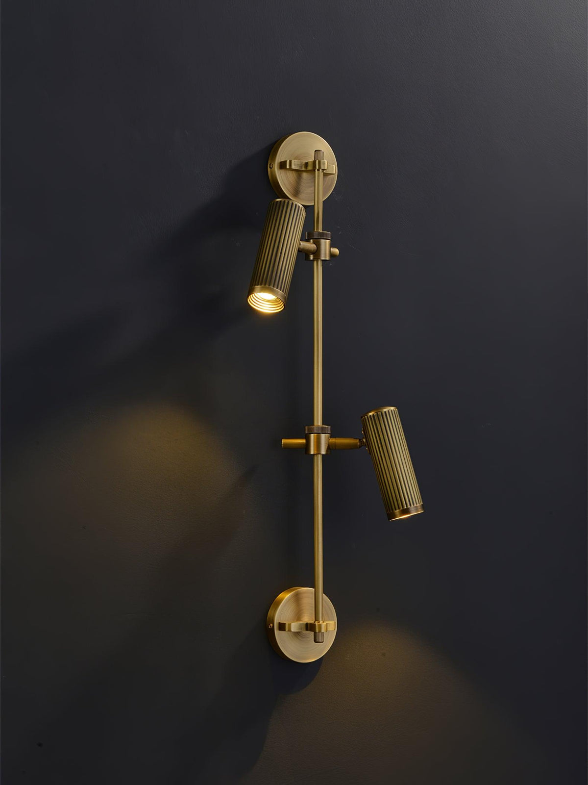 Two-head Clérachic wall light on a dark wall, with the upper head lit and aimed down and the lower head angled out.