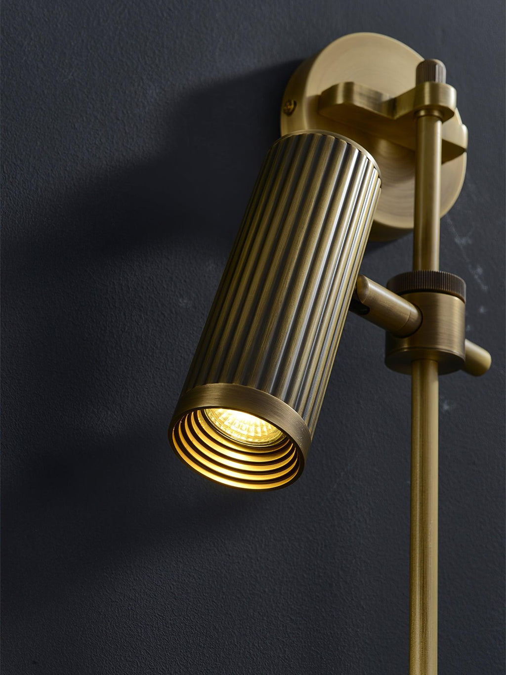 Fluted bronze head lit from inside its ringed lens, close up on a dark wall, from this Clérachic wall light.