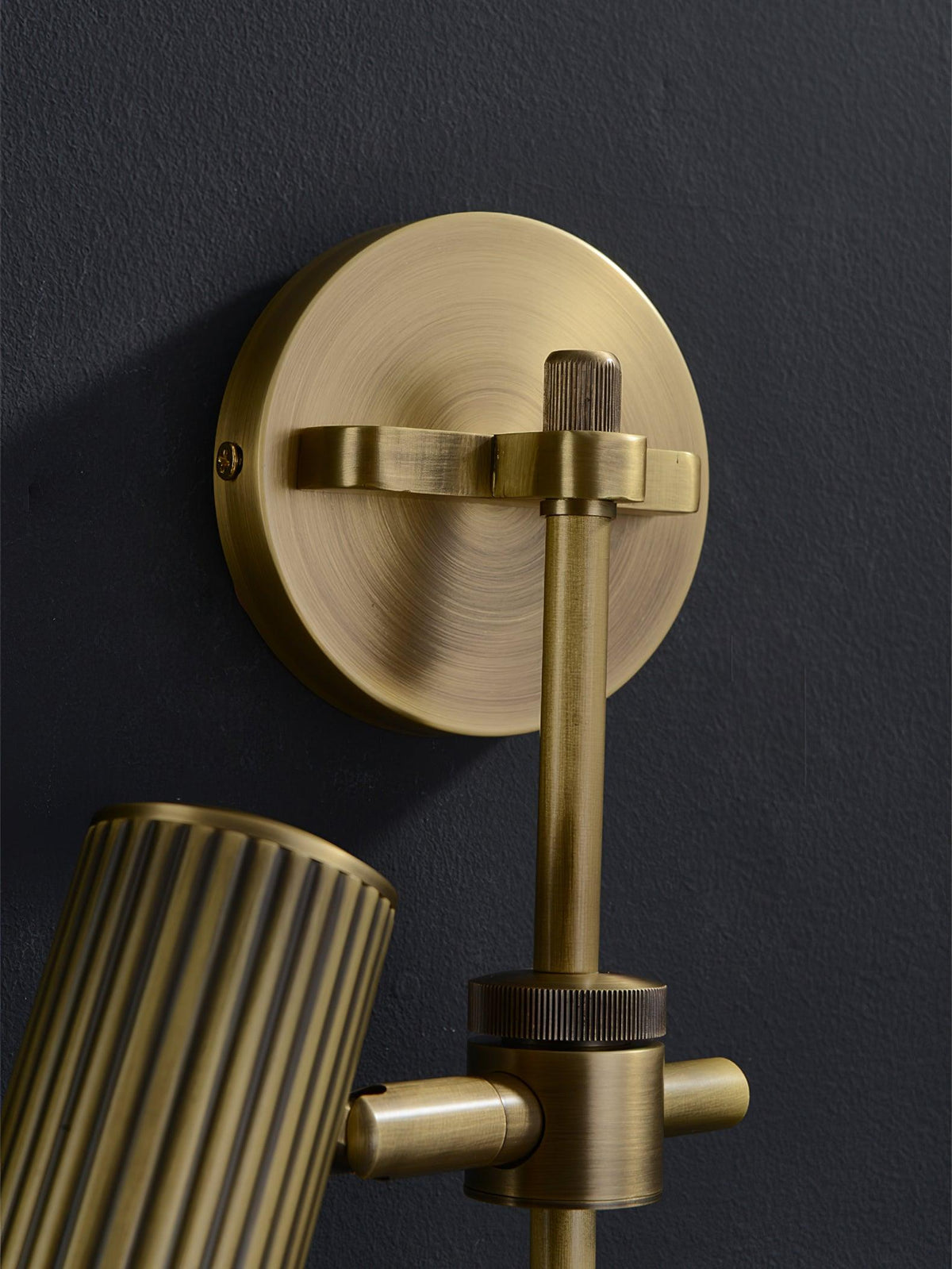 Round bronze backplate, rail and pivot joint of this Clérachic wall light, with part of one fluted head, against a dark wall.