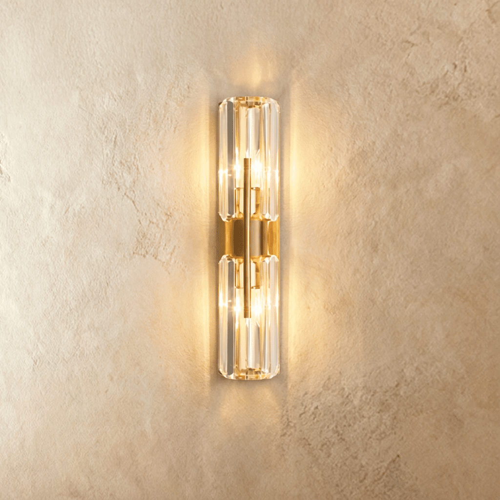 Clérachic brass wall lamp with two stacked faceted crystal cylinders, lit warm on a limewash wall.