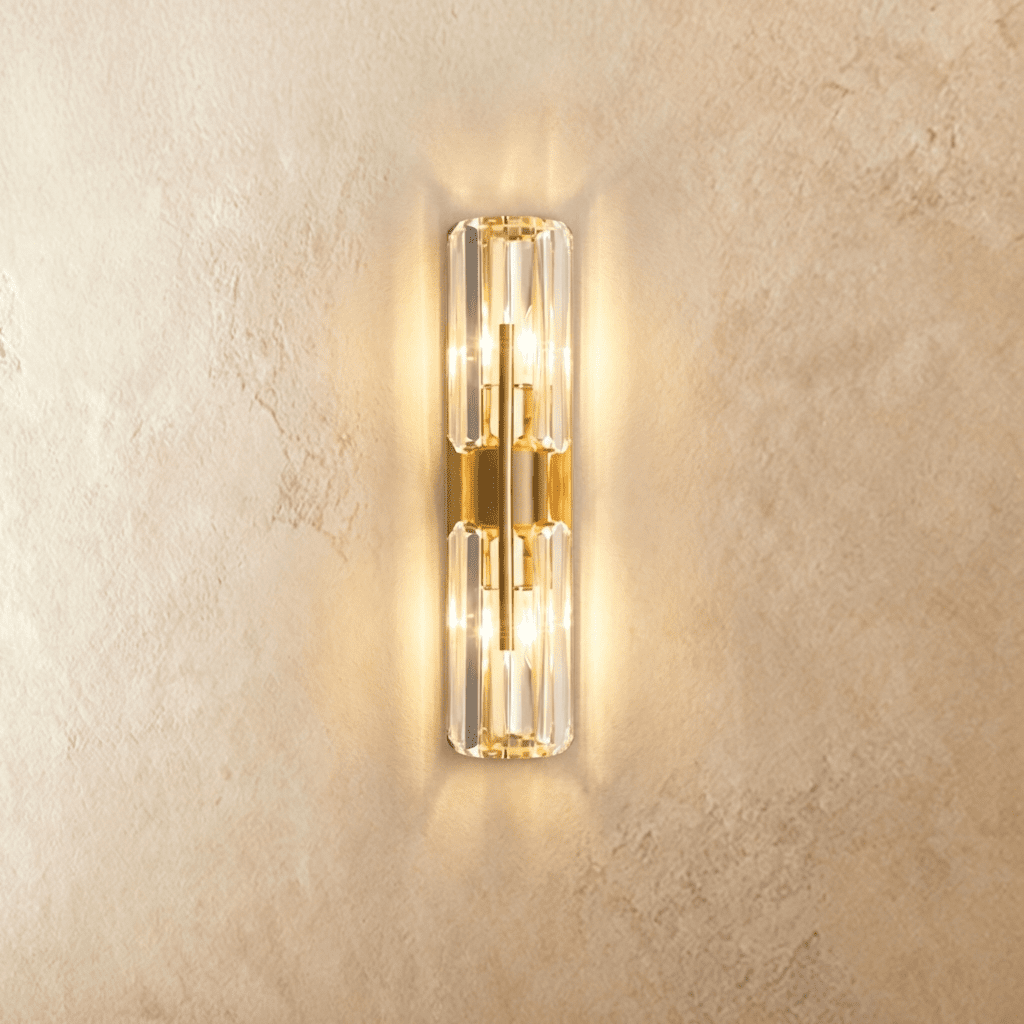 Clérachic wall lamp with two faceted crystal cylinders on a central brass column, glowing warm against a textured wall.