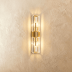 Clérachic wall lamp with two faceted crystal cylinders on a central brass column, glowing warm against a textured wall.