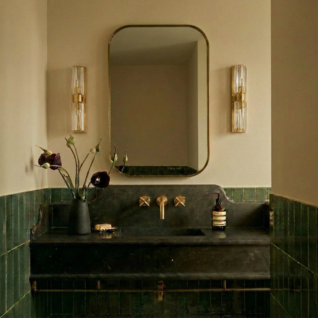 Pair of Clérachic brass and crystal wall lamps flanking a rounded brass-framed mirror above a dark stone basin and green tiles.