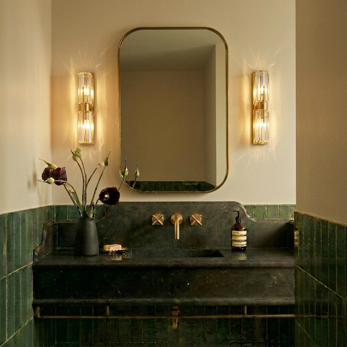 Pair of Clérachic crystal wall lamps lit warm beside a brass-framed mirror in a bathroom with green tiles and brass taps.