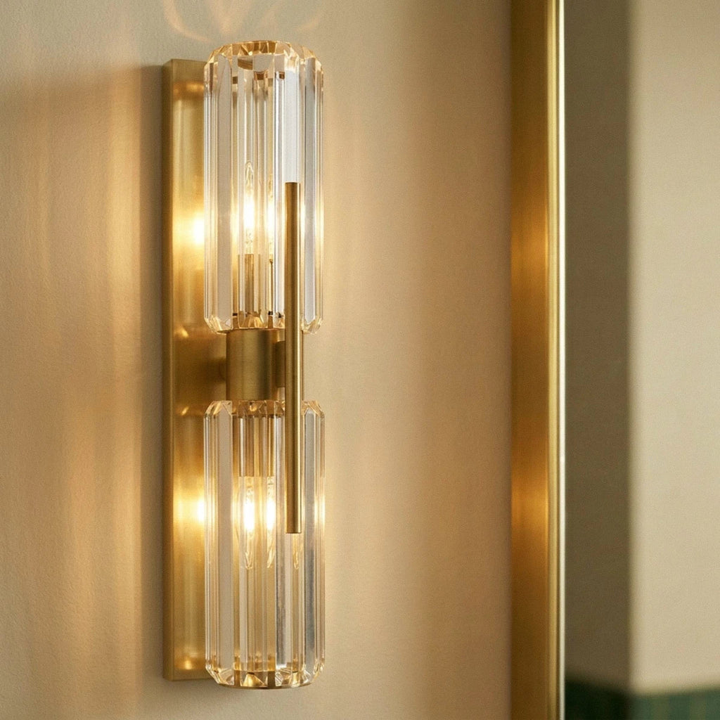 Close view of a Clérachic brushed brass wall lamp with two ribbed crystal cylinders lit warm beside a brass mirror frame.