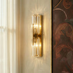 Clérachic brass wall lamp with two faceted crystal cylinders casting patterned light on a plaster wall beside a sheer curtain.