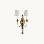 Two pleated white shades on antique brass arms and a black tapered column, this Clérachic wall light on a plain white backdrop.