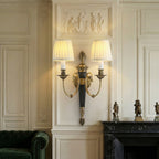 Lit Clérachic wall light with two pleated shades on a panelled cream wall, above a black marble fireplace and a green velvet sofa.