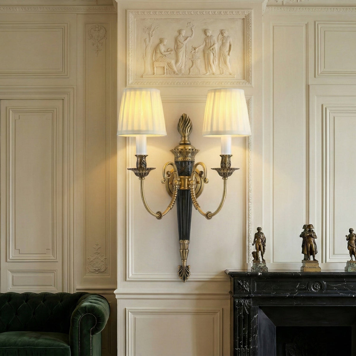 Lit Clérachic wall light with two pleated shades on a panelled cream wall, above a black marble fireplace and a green velvet sofa.