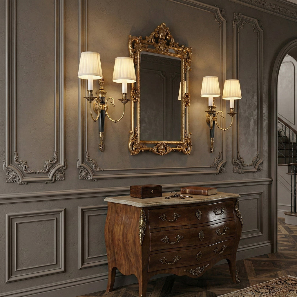 A pair of two-arm wall lights flank a gilt mirror above a walnut chest of drawers, one a Clérachic piece, on a grey panelled wall.