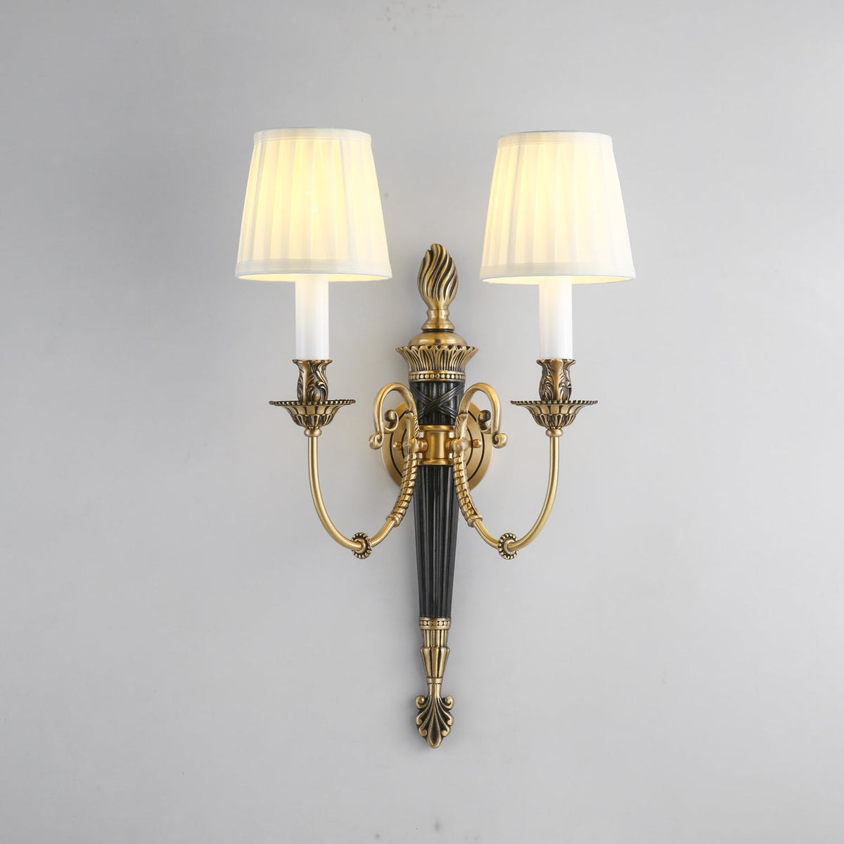 Front view of this Clérachic wall light on a pale grey wall, showing two lit pleated shades, a flame-shaped top and curved brass arms.