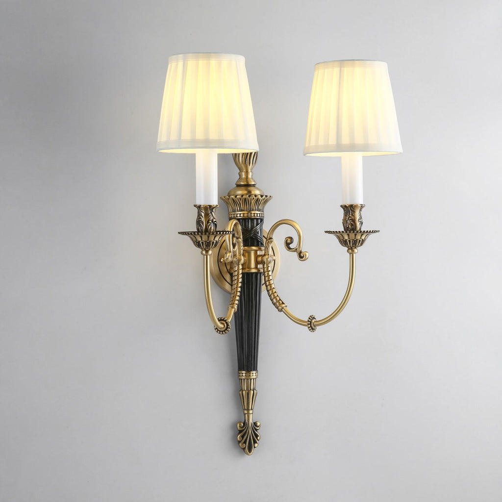 Slightly angled view of this Clérachic wall light, its pleated shades glowing warm above scrolled brass arms and a black column.