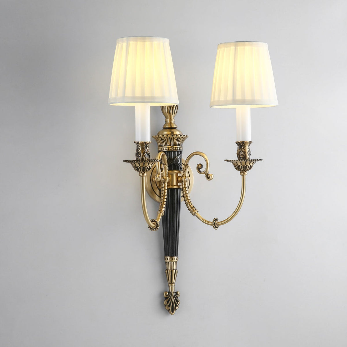 Slightly angled view of this Clérachic wall light, its pleated shades glowing warm above scrolled brass arms and a black column.