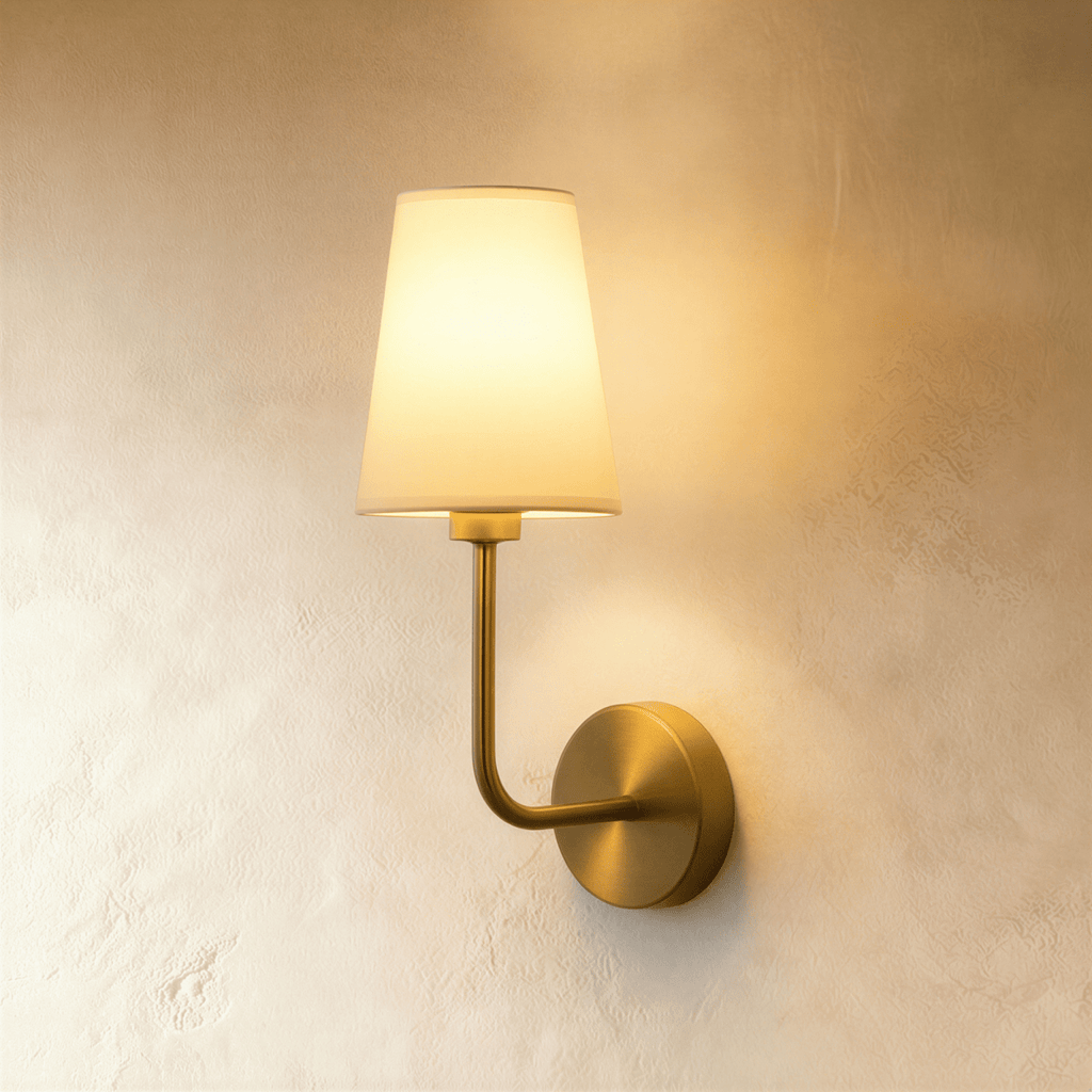Clérachic wall lamp with a round brushed-brass backplate, curved brass arm and off-white fabric shade, glowing warm on plaster.