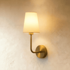 Clérachic wall lamp with a round brushed-brass backplate, curved brass arm and off-white fabric shade, glowing warm on plaster.