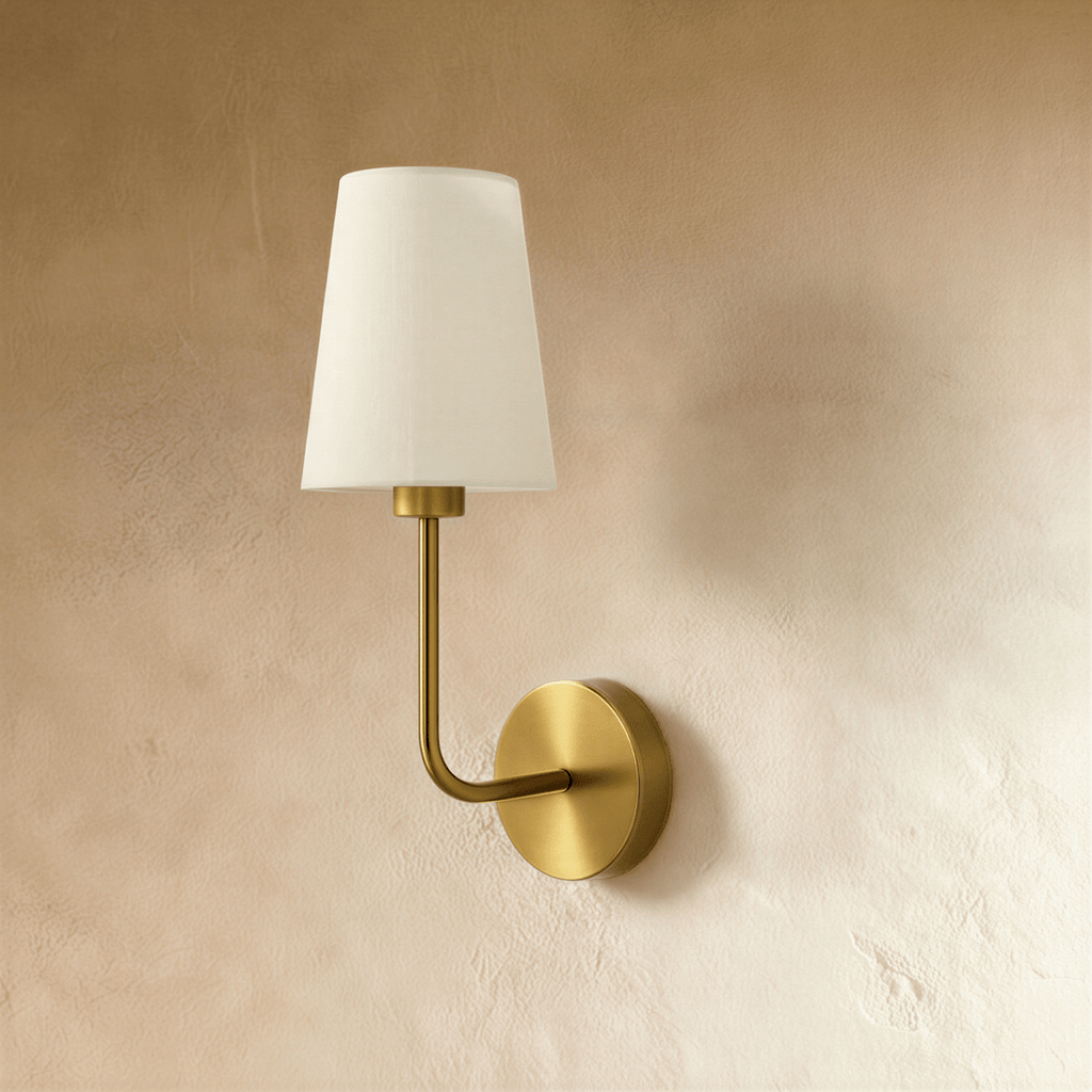 Clérachic wall lamp in brushed brass with a round backplate, an elongated curved arm and a tapered off-white fabric shade, unlit.