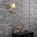 Clérachic brass wall lamp with an off-white shade, lit warm against grey stacked-stone panelling above a black cabinet.