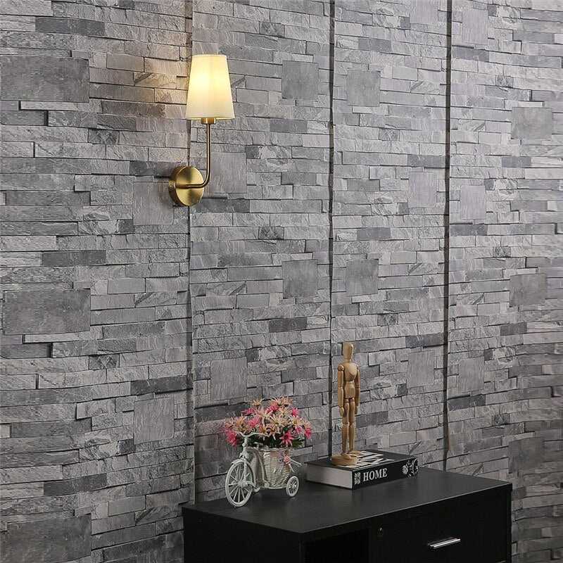 Clérachic brass wall lamp with an off-white shade, lit warm against grey stacked-stone panelling above a black cabinet.
