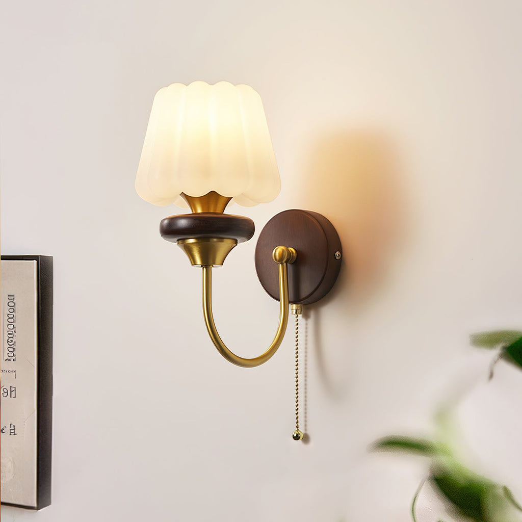 Lit Clérachic wall light with a fluted white glass shade and a curved brass arm, on a pale wall beside a framed print and a plant.