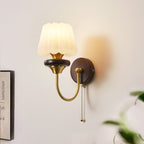Lit Clérachic wall light with a fluted white glass shade and a curved brass arm, on a pale wall beside a framed print and a plant.