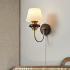 A Clérachic wall lamp with a tapered white glass shade and walnut ring, on a grey wall above a woven-backed headboard.