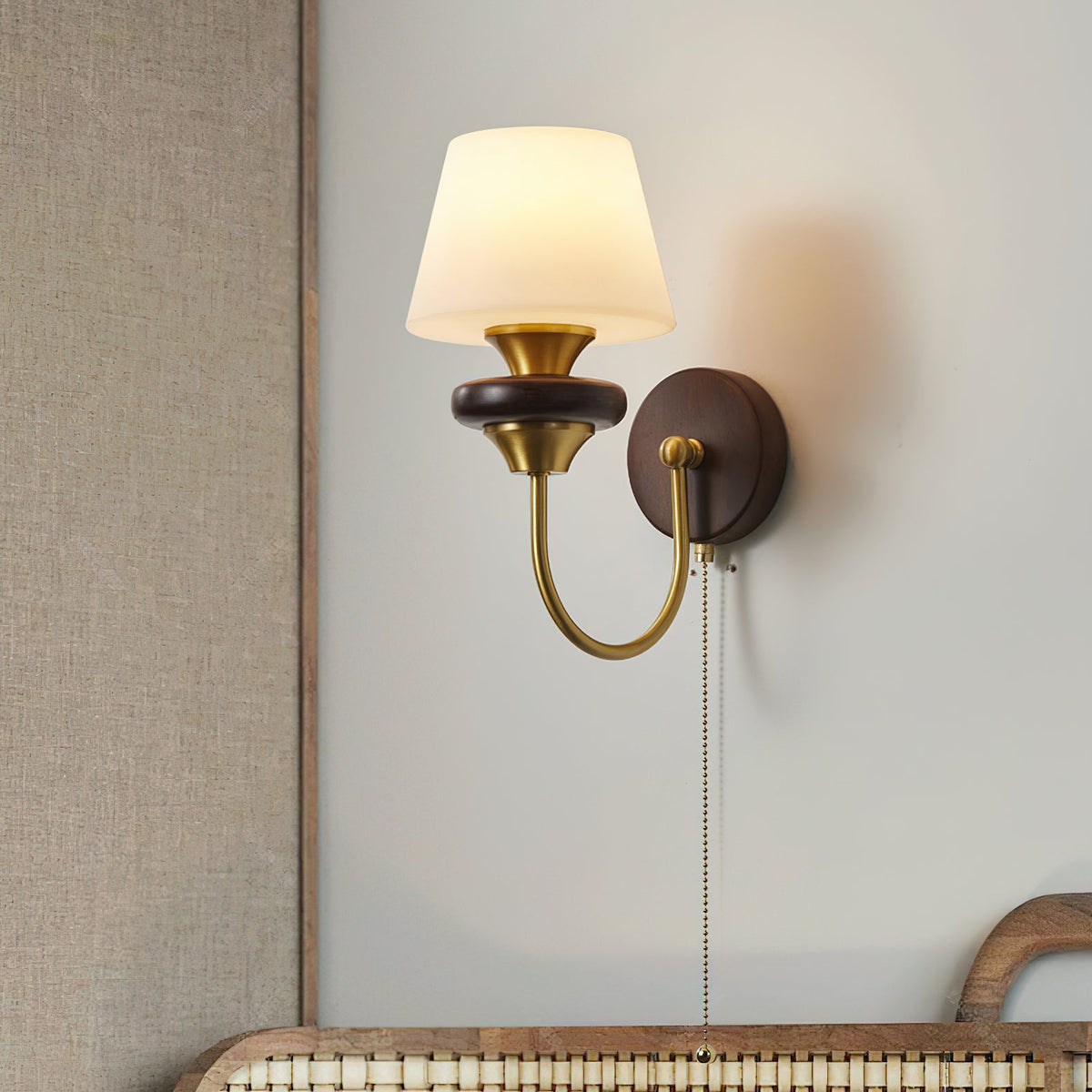 A Clérachic wall lamp with a tapered white glass shade and walnut ring, on a grey wall above a woven-backed headboard.