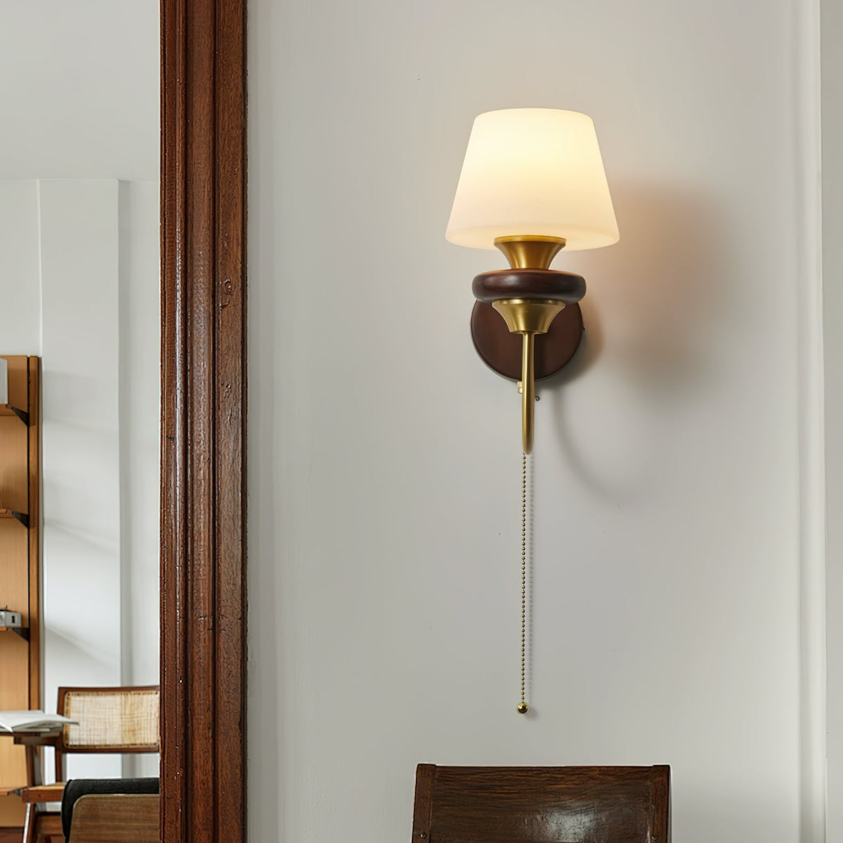 A tapered white-shade Clérachic wall light on a straight brass arm, with a long pull chain, beside a dark wood door frame.