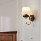 A fluted white glass Clérachic wall lamp on a curved brass arm, glowing warm on a pale wall above a dark wood chest.