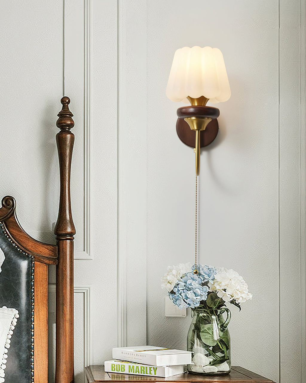 A Clérachic fluted-shade wall light above a bedside table with hydrangeas and books, beside a carved wooden bedpost.