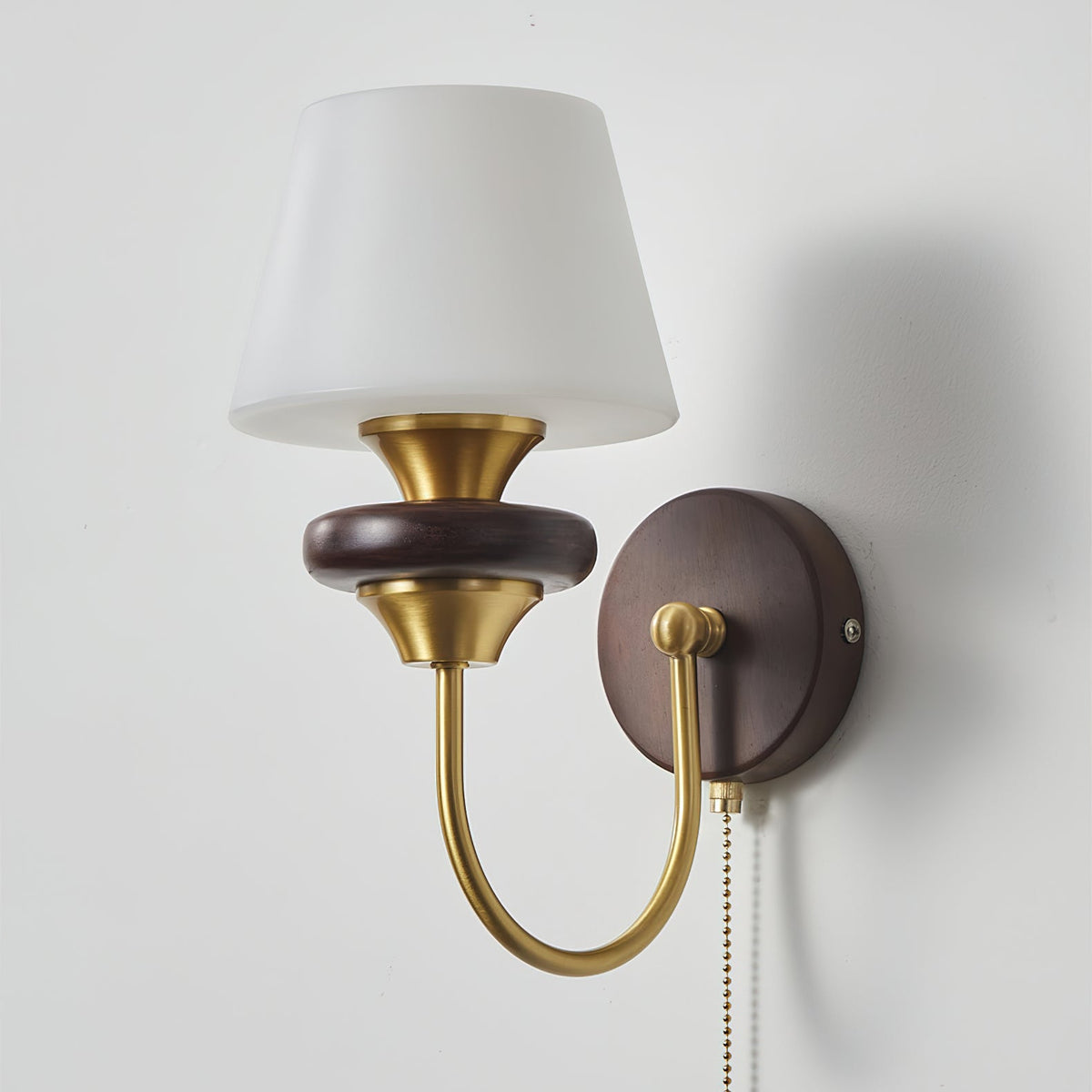 Close view of this Clérachic wall lamp with a tapered white glass shade, a brass cup, a walnut ring and a curved brass arm.