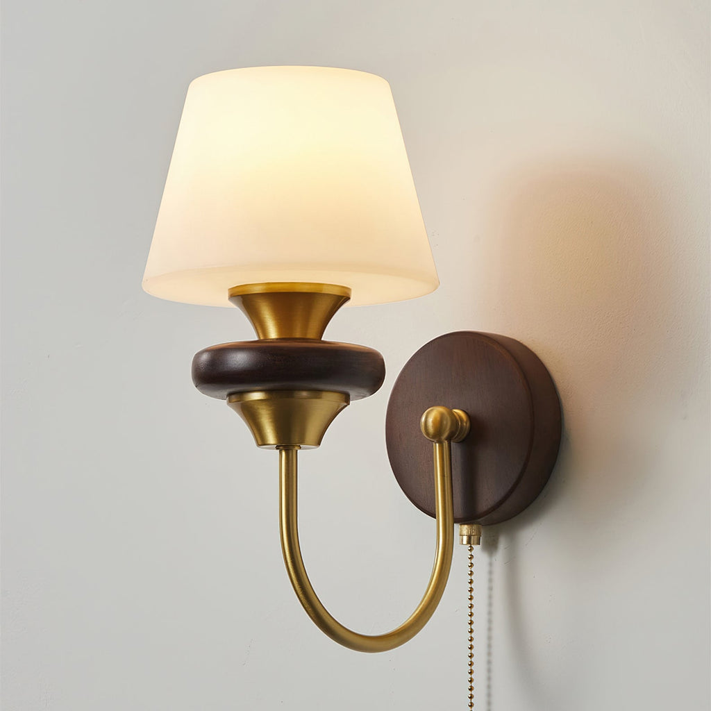 A lit Clérachic wall lamp with a tapered frosted glass shade, a brass arm and a walnut-toned round backplate.