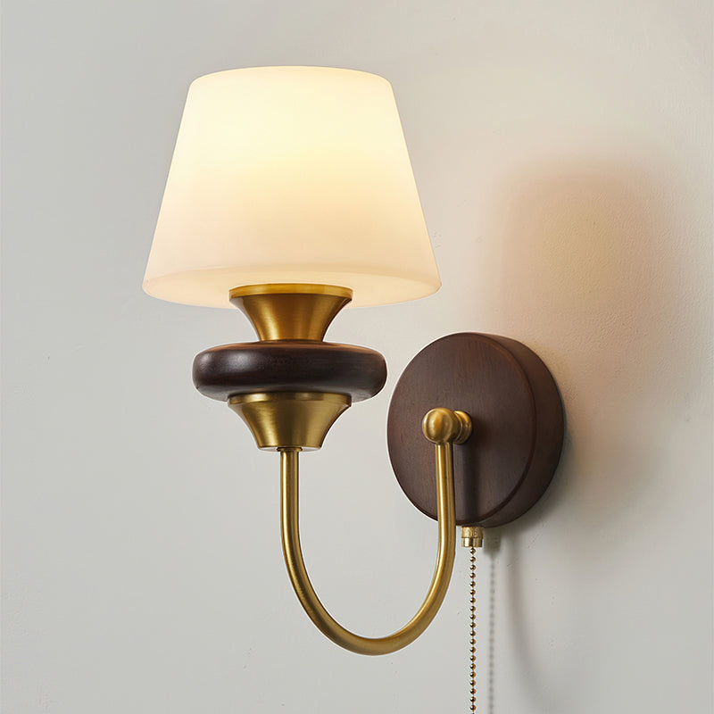 A Clérachic wall lamp with a tapered frosted glass shade, lit warm, on a curved brass arm beside a round walnut backplate.