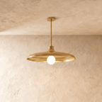 Clérachic pendant light in a brighter brass finish, its wide disc shade and opal glass bulb below a plaster ceiling.