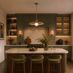 Clérachic brass pendant light glowing above a marble kitchen island with green cabinetry and velvet barstools.