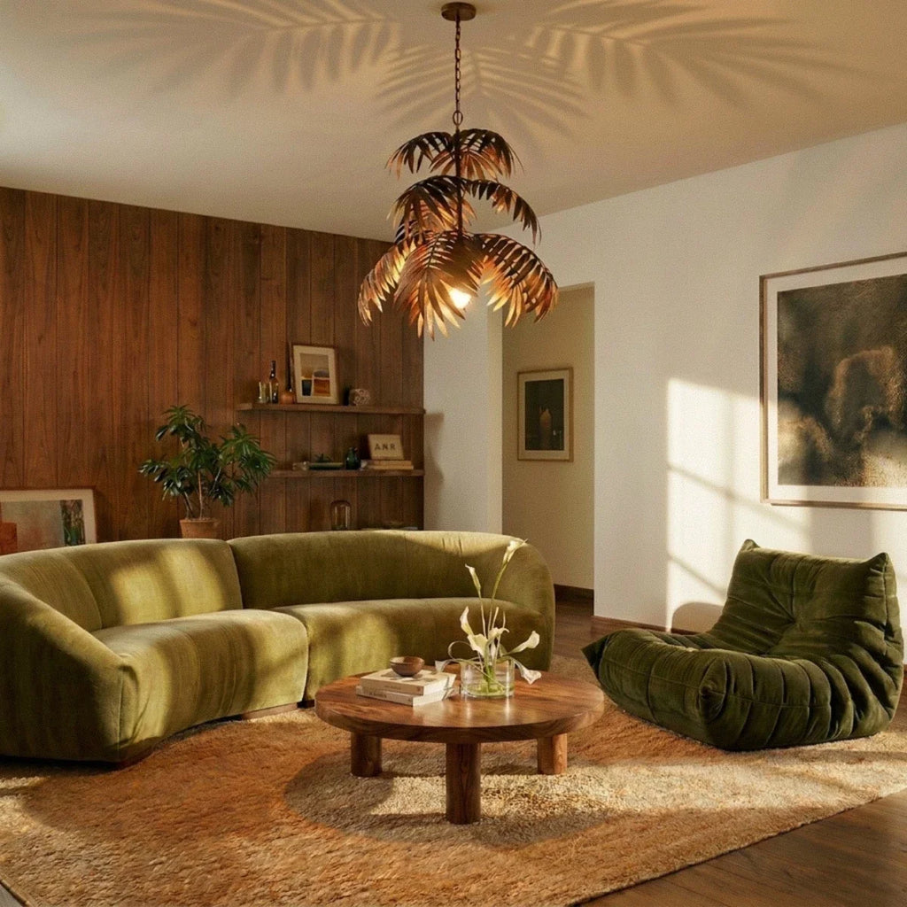 Clérachic tiered bronze palm-leaf chandelier above a curved olive sofa and round wooden table, casting leaf shadows on the ceiling.