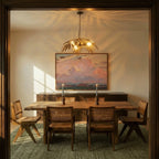 Clérachic hanging palm-leaf chandelier in antique gold, lit above a rustic wooden dining table and cane-backed chairs.
