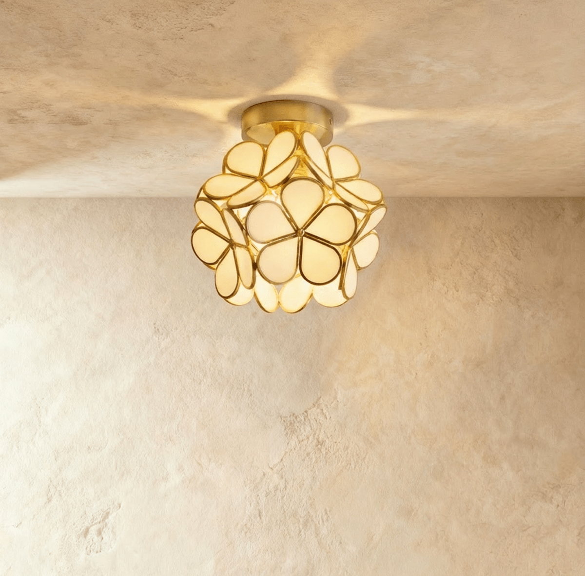 Clérachic flower ceiling light in cream glass petals on a brass mount, glowing warm on a plaster ceiling.