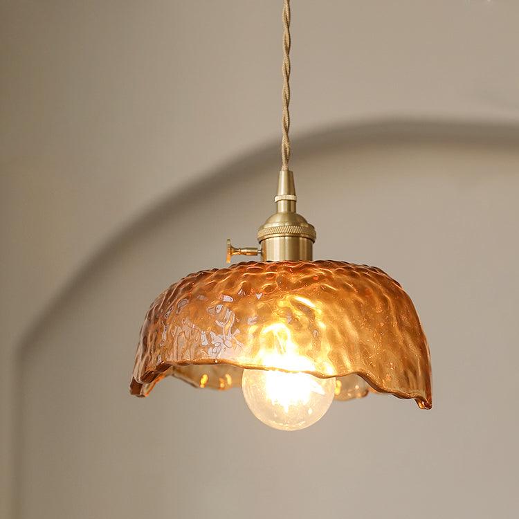 Lit Clérachic pendant with a rippled amber-brown glass dome, a brass socket with a small switch, and a round bulb glowing against a warm beige wall.