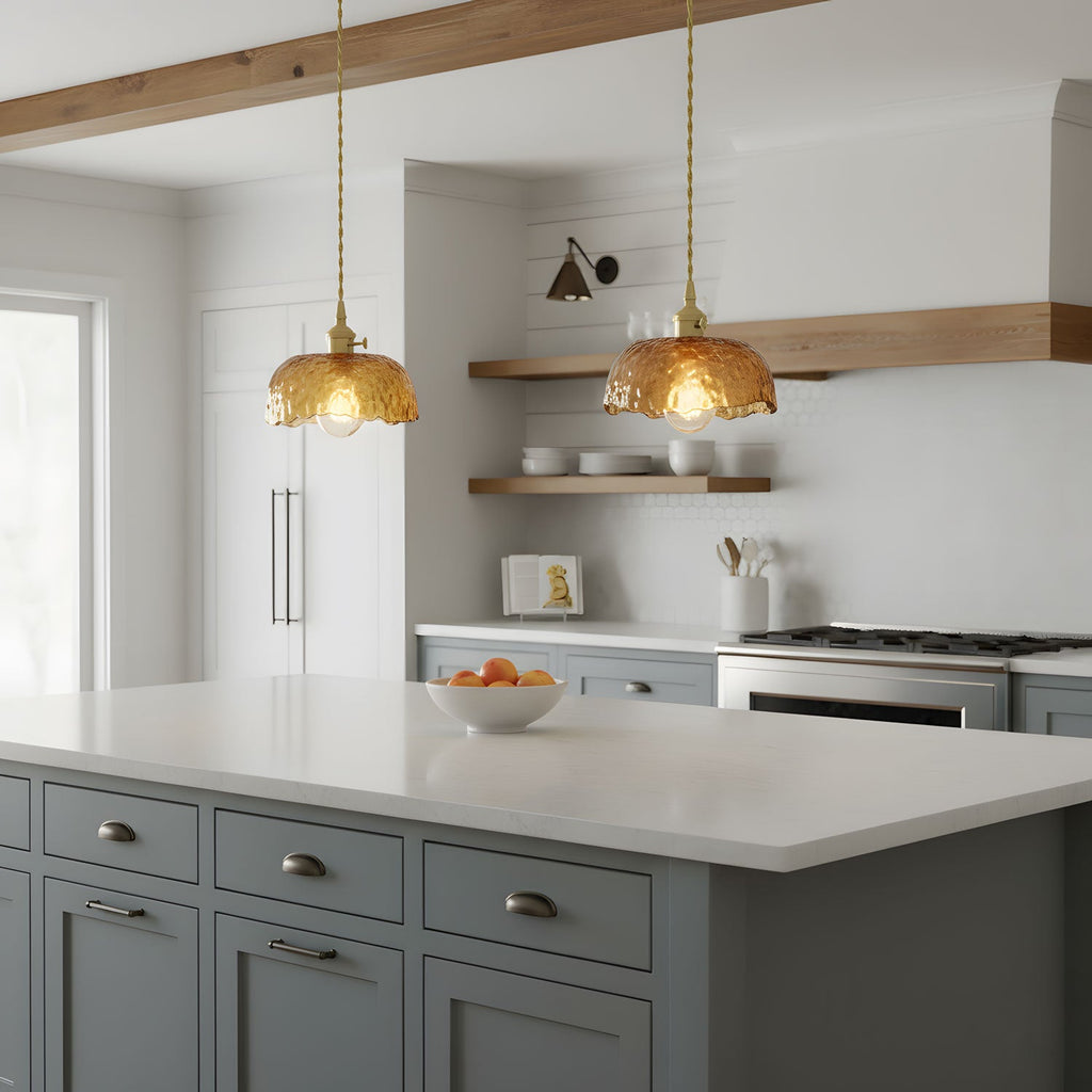 Two Clérachic brown glass pendants with scalloped domes over a white kitchen island with grey-blue drawers and open wooden shelves behind.