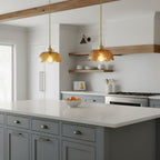 Two Clérachic brown glass pendants with scalloped domes over a white kitchen island with grey-blue drawers and open wooden shelves behind.