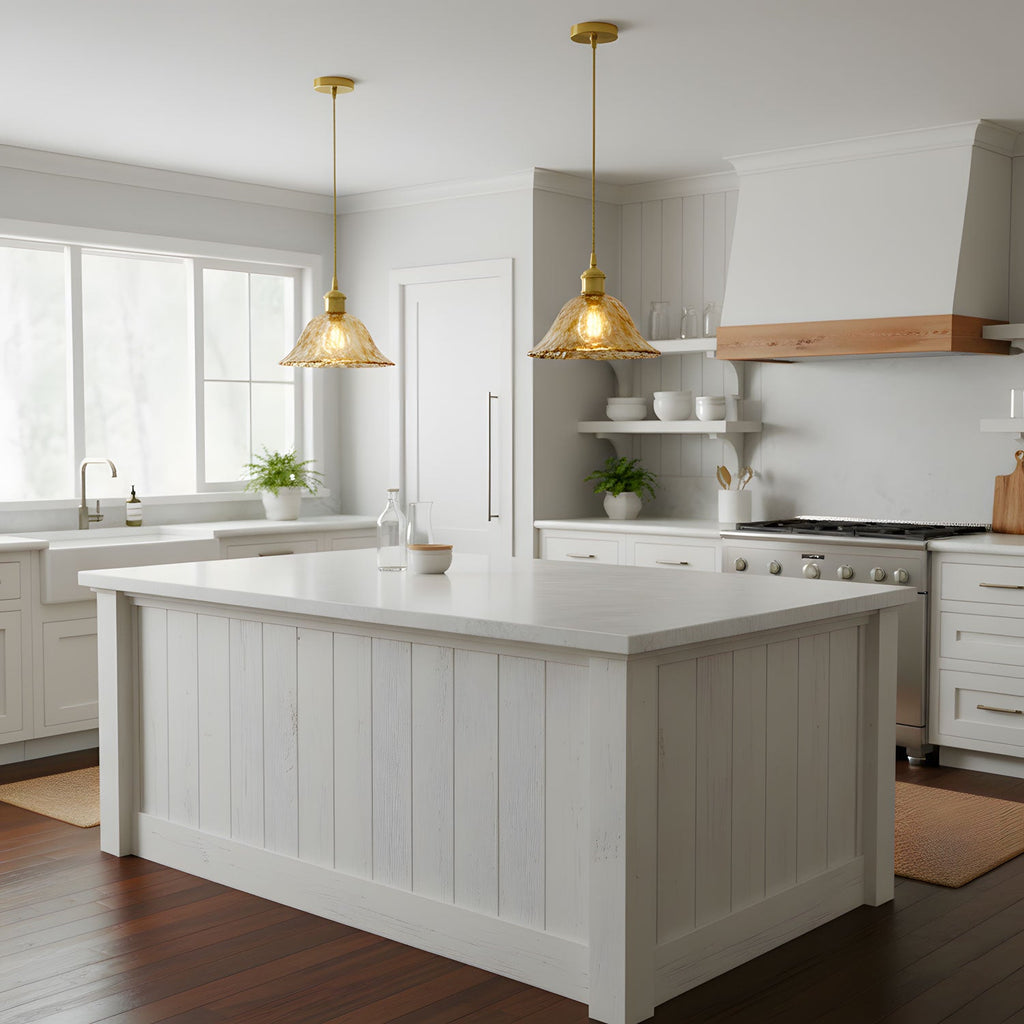 Two brass-topped Clérachic glass pendants glowing over a white panelled kitchen island, with a pale range hood and a window behind.