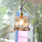 Clérachic cube-shaped brown glass pendant glowing outdoors against blurred green leaves, hung from a dark twisted cord and a brass socket.
