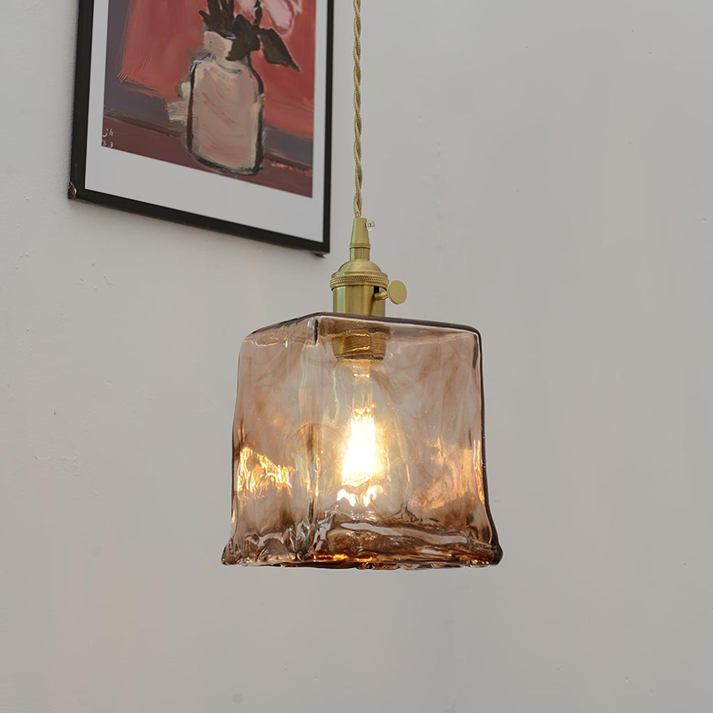 This Clérachic cube pendant in swirled brown glass, with a filament bulb lit, hanging on a grey wall beside a framed still life.