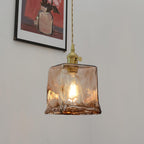 This Clérachic cube pendant in swirled brown glass, with a filament bulb lit, hanging on a grey wall beside a framed still life.